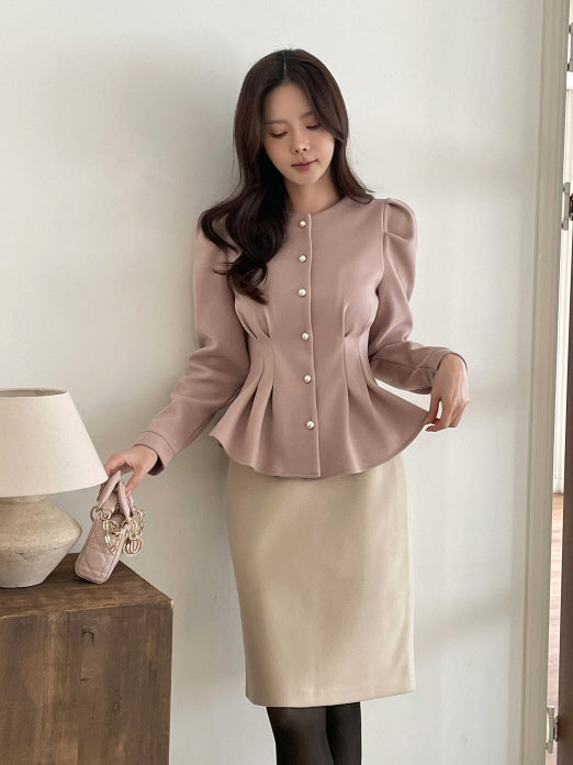淡いピンクのパフショルダージャケットとベージュスカートを合わせた女性が室内で優しくポーズを取る全身コーデ。パールボタンとペプラムシルエットが大人可愛いオフィスカジュアルの雰囲気を演出。