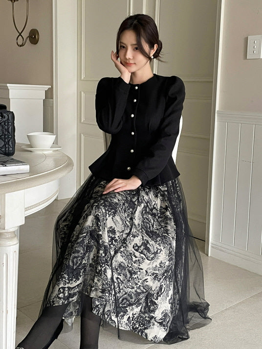 ブラックのペプラムジャケットにモノトーン柄のロングスカートを合わせ、椅子に座って微笑む女性。パールボタンとふんわりショルダーがクラシックなエレガンスを感じさせる大人フェミニンコーデ。