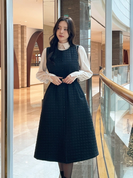 アイボリーブラウスとキルティングワンピースを重ねた女性がガラス手すりの前に立つシーン。ハリのあるスクエアキルティングが美しいAラインを作り、大人きれいめな冬のレイヤードコーデに仕上がっている