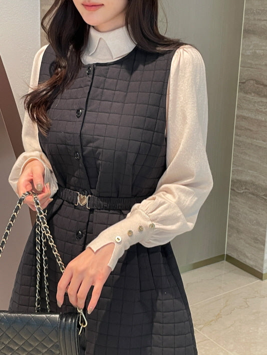 アイボリーのブラウスに黒のキルティングワンピースを重ねた女性がバッグを持つ上半身のスタイル。立体的なスクエアキルティングとハートベルトが上品な大人フェミニンコーデを引き立てる。