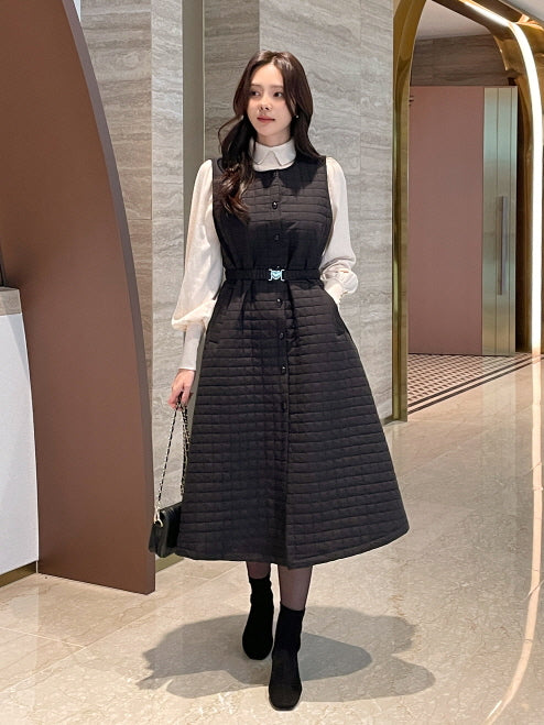 黒のキルティングAラインワンピースにブラウスを合わせた女性がホテルの廊下でポーズを取る全身コーデ。ふんわり広がるシルエットとレイヤードスタイルがクラシックな上品さを演出。