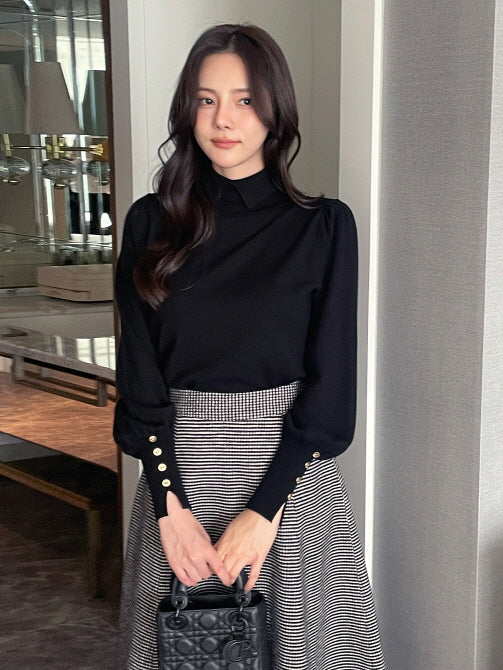 黒の襟付きトップスを着た女性が、袖口のゴールドボタンをポイントに千鳥格子のフレアスカートと合わせた上品フェミニンなオフィスカジュアルコーデ